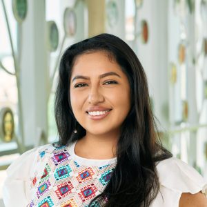 A latina woman smiling, with long brown hair, wearing a white top with geometric pattern and dangly pearl earrings.